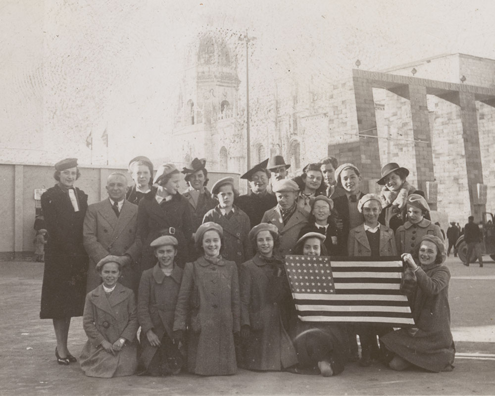 A photo of a group of girls in identical grey berets and overcoats standing in front of a monastery, holding an American flag. In the back row there are adults in black overcoats and varying hats. 