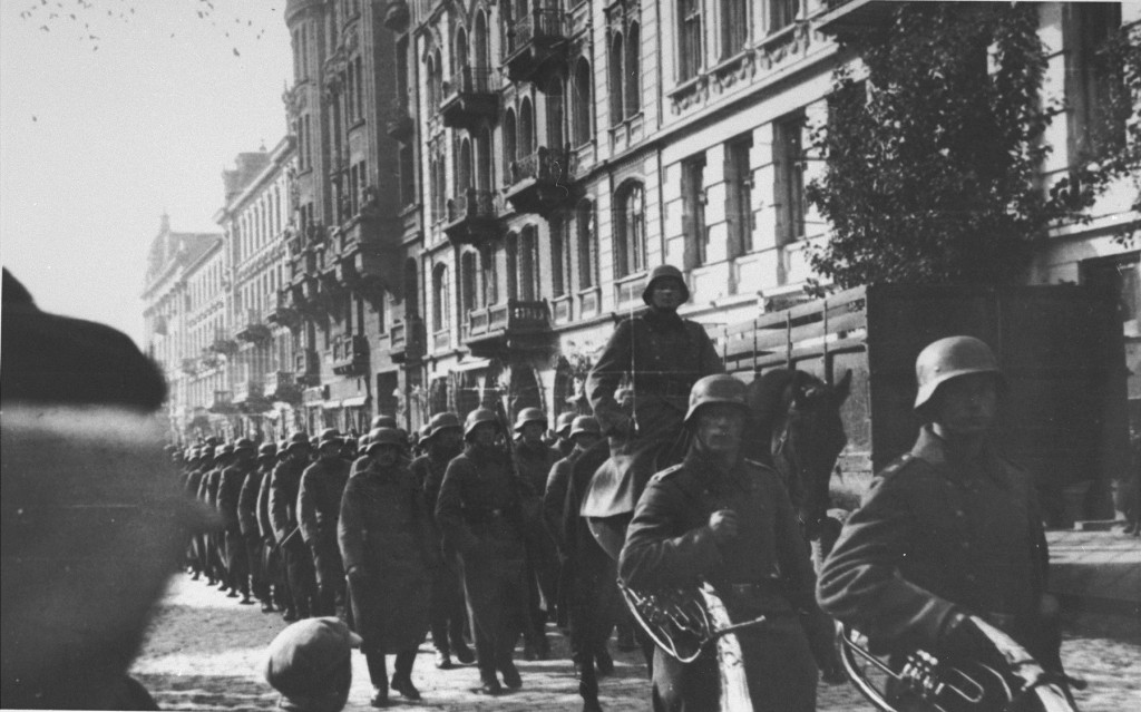 Rows of soldiers parade down the street, wearing Nazi-era helmets. Curiously, the two in the front are carrying tubas. 