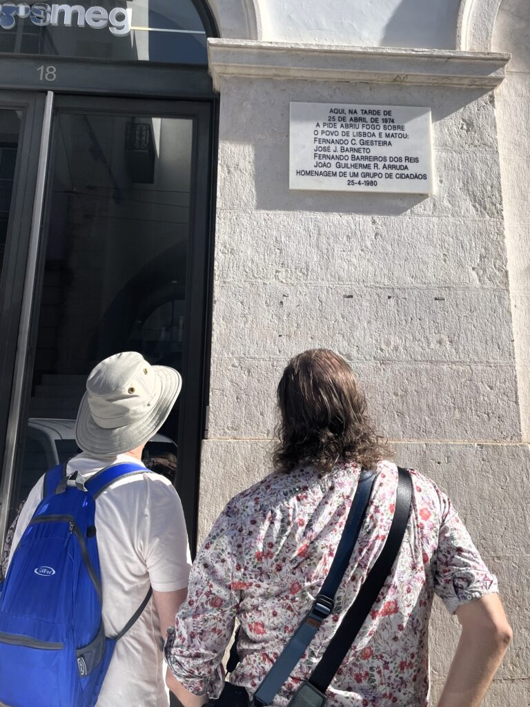 pide-revolution-tour Two men stand in front of a plain white building with a simple plaque. That plaque lists the names of the four people who were killed there when the secret police opened fire on April 25, 1974.